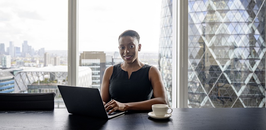 A female executive works at her desk in a skyscraper, with 30 St Mary Axe (the Gherkin) visible through the floor to ceiling windows.