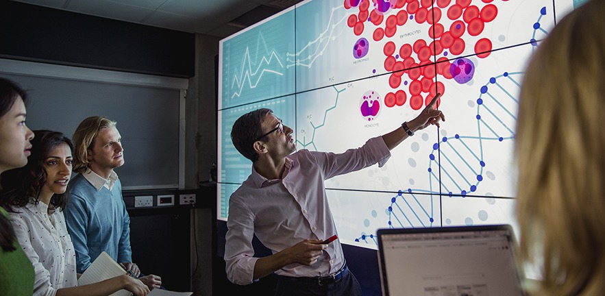 Group of business professionals in a dark room standing in front of a large data display screen with information.