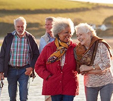 Group of smiling senior friends walking arm-in-arm along shoreline of winter beach.