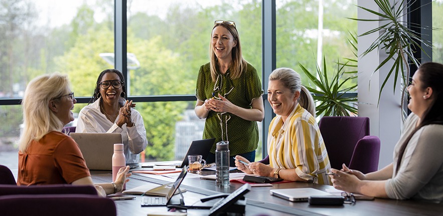 A wide-angle view of a group of women laughing and enjoying listening to each other's ideas in a business meeting.