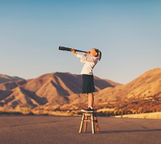 A young student dressed in business attire stands on a stool looking through a telescope.