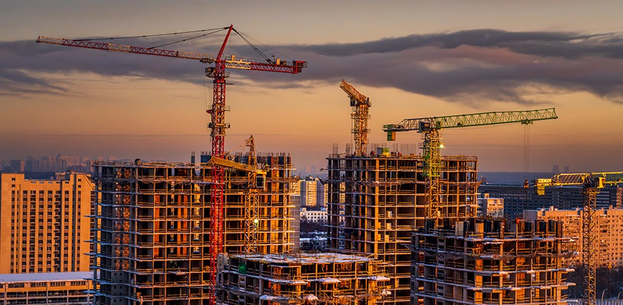 Multiple unfinished apartment buildings with visible concrete frames and tower cranes in a dense urban area during evening light. Image is suitable for illustrating residential development, construction industry, or urban growth.