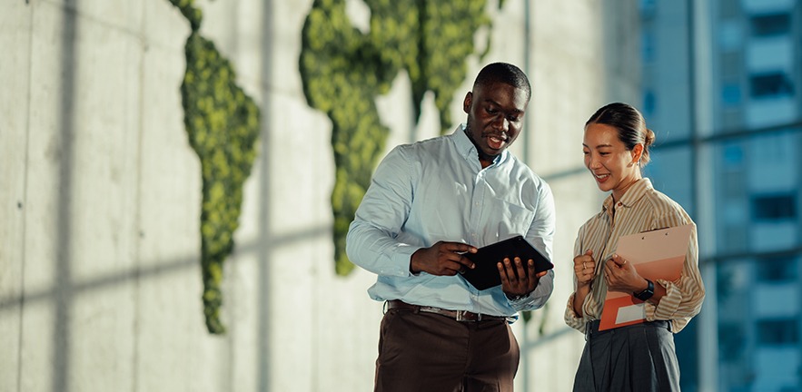 Two managers are discussing sustainable business strategies using digital tablet and clipboard, with a green world map in the background symbolising global environmental awareness.
