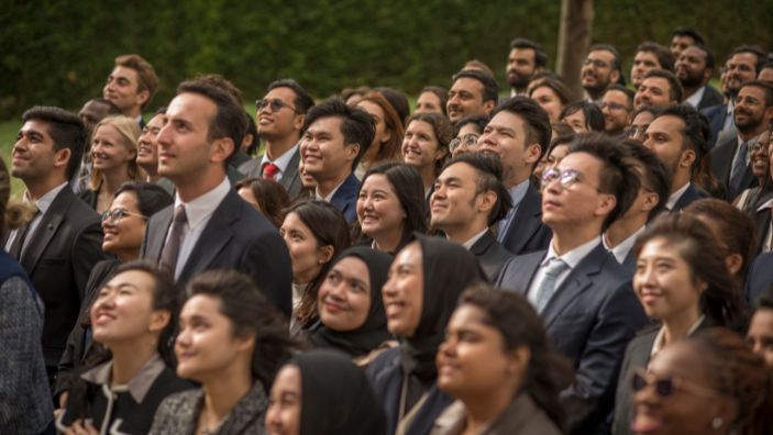 MBA students all looking the same direction and smiling outside, possibly listening to a speech.