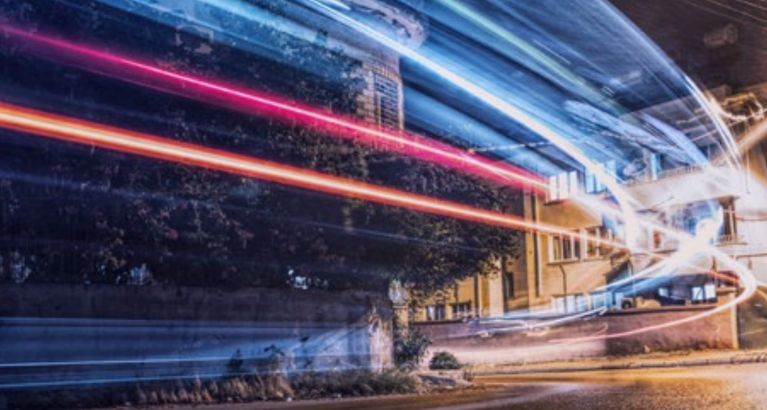 Long-exposure light trails passing through a city street at night, with buildings and trees in the background.