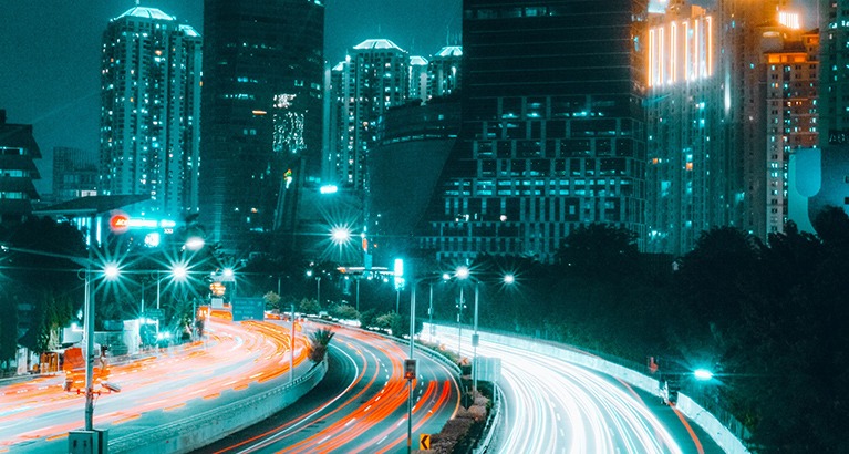 Long-exposure light trails passing through a city street at night, with buildings and trees in the background.