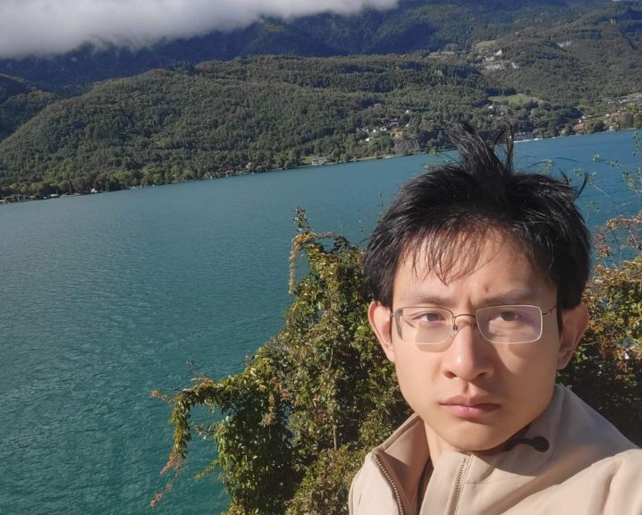 Hanzhe Xing in front of a lake with trees, hills and dramatic clouds in the background.