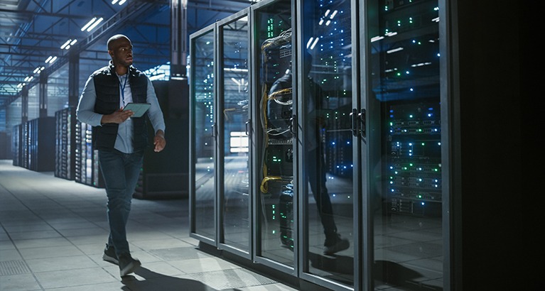 Data center IT specialist walks through aisles of server racks, using a tablet to inspect equipment, monitor network activity, and check connections in a cyber secure server room.